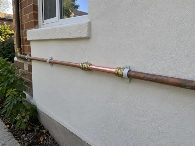 Exterior copper water pipe repair on a rendered house wall with window and brick corner in Croydon