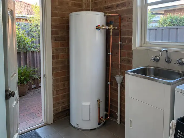 White hot water cylinder beside a laundry sink and brick wall with copper pipes in Ashfield